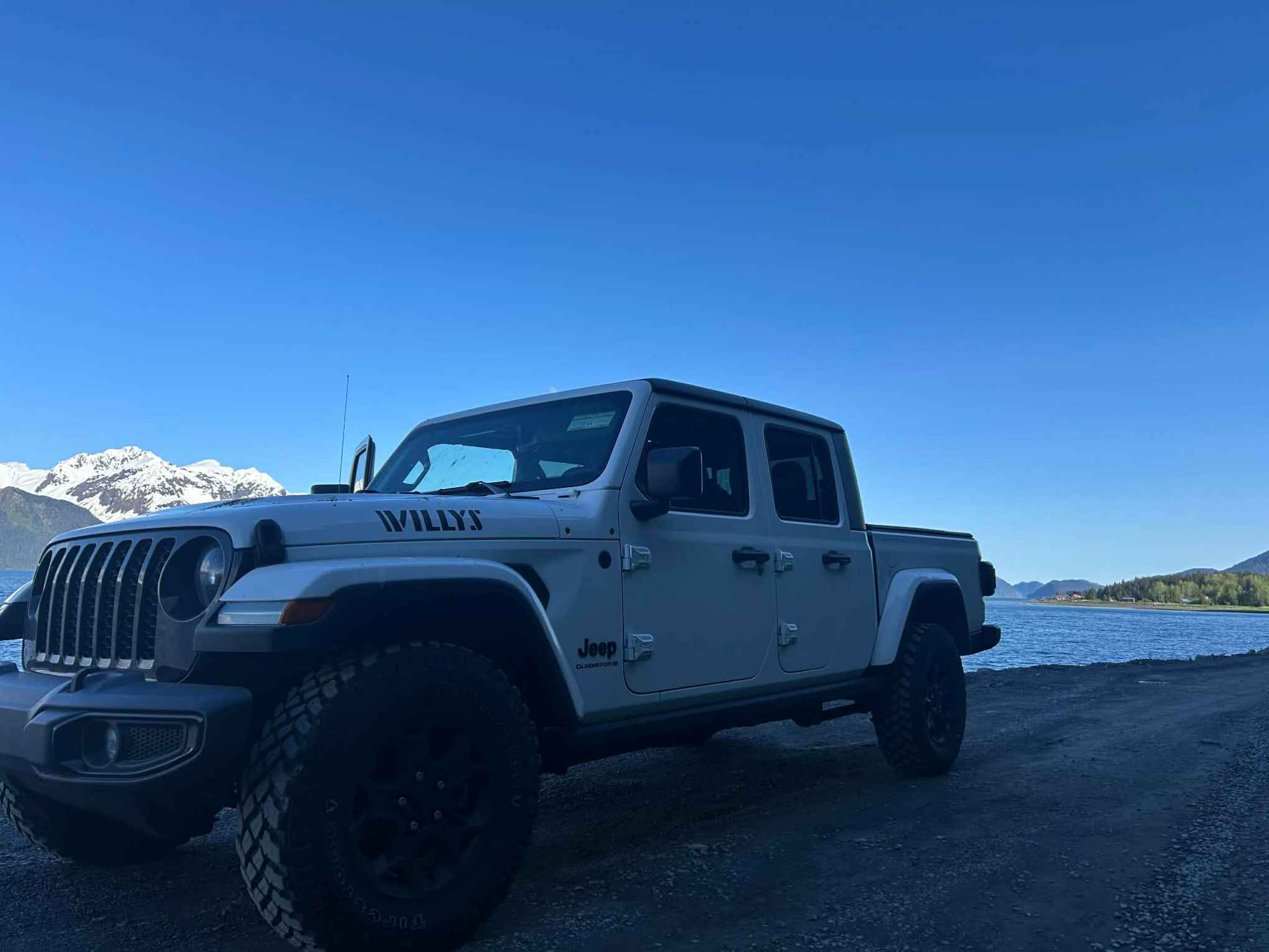 White Jeep parked near a lake with snow-capped mountains in the background.