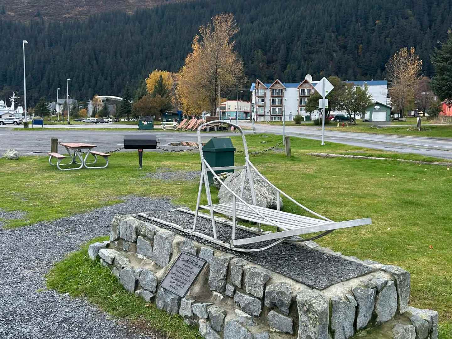 Outdoor sled on stone platform with plaque, grassy area, and trees; buildings and hills in background.