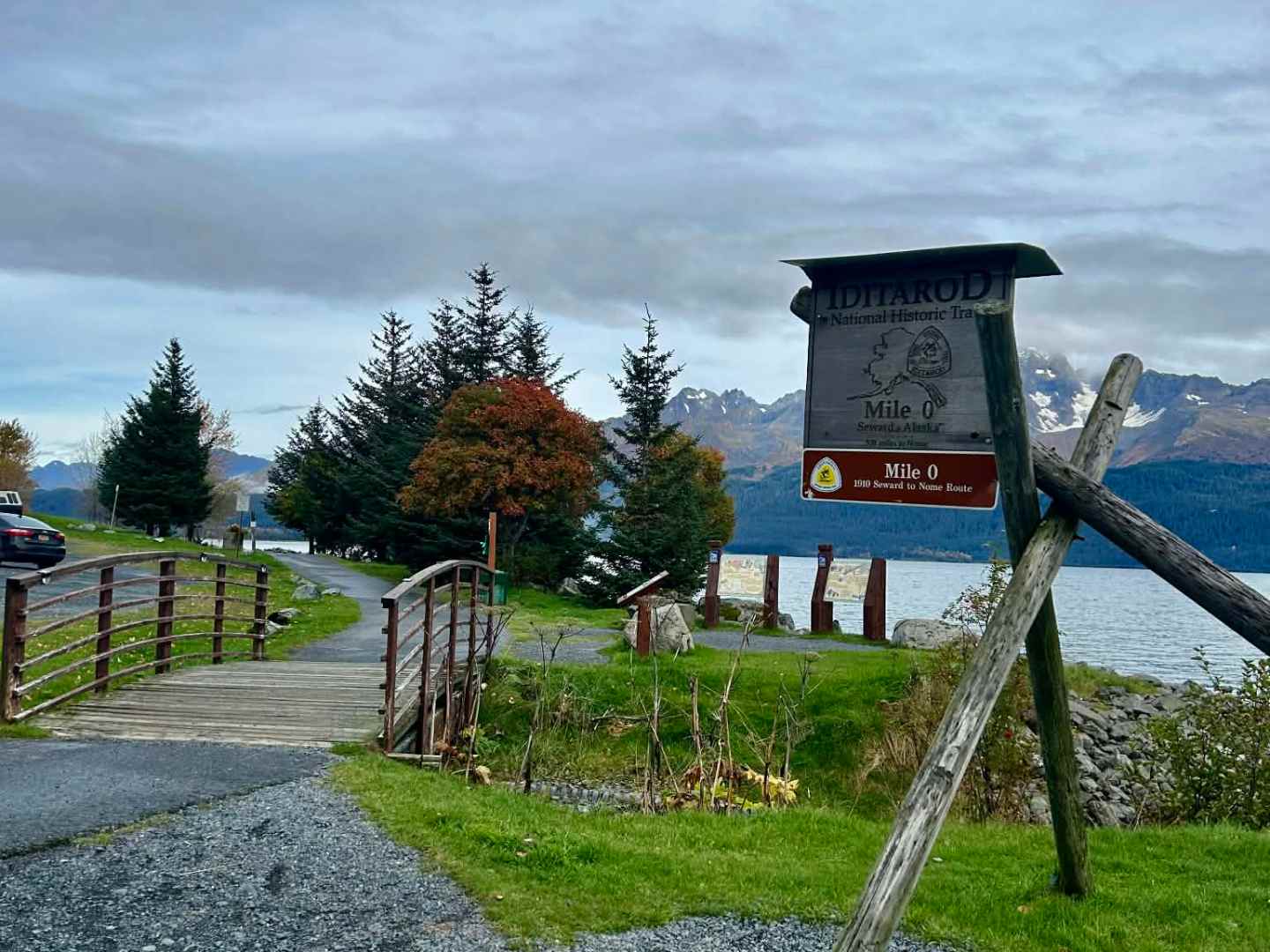 Iditarod Mile 0 sign by a small path with mountains and trees in the background.