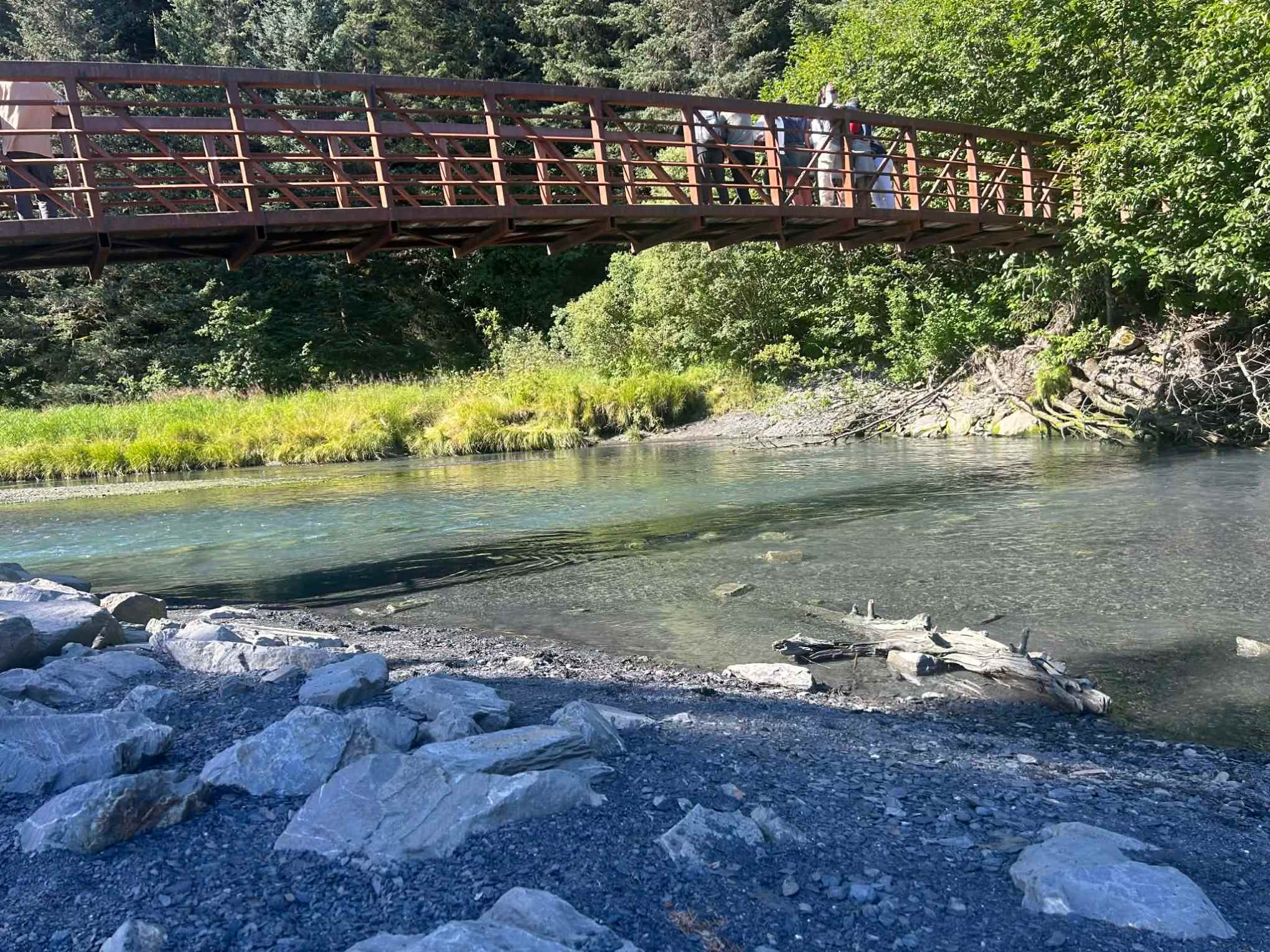 Bridge over a clear stream with rocky banks and lush greenery in the background.