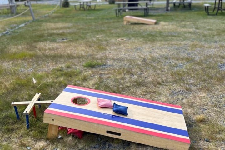 Cornhole board with bean bags on grass, tents, and mountains in the background.