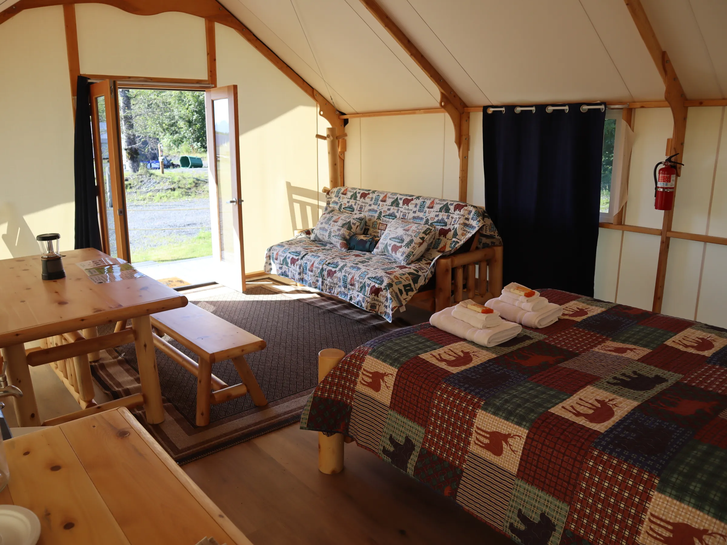 Glamping cabin interior with a bed, sofa, and wooden dining set, open door to outdoors.