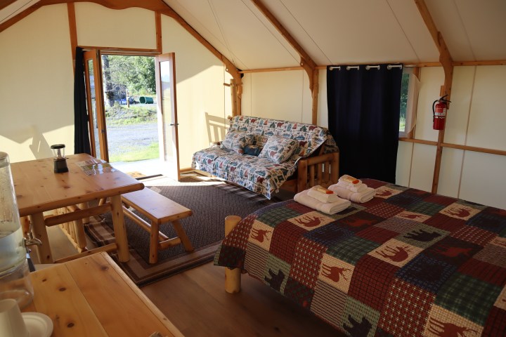 Glamping cabin interior with a bed, sofa, and wooden dining set, open door to outdoors.