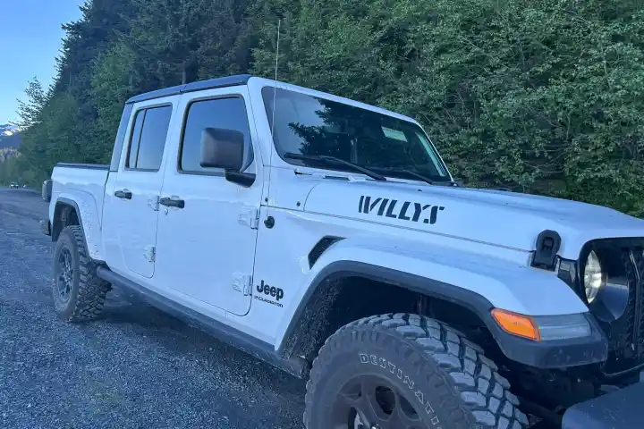 White Jeep Gladiator Willys parked on gravel with trees and mountains in the background.