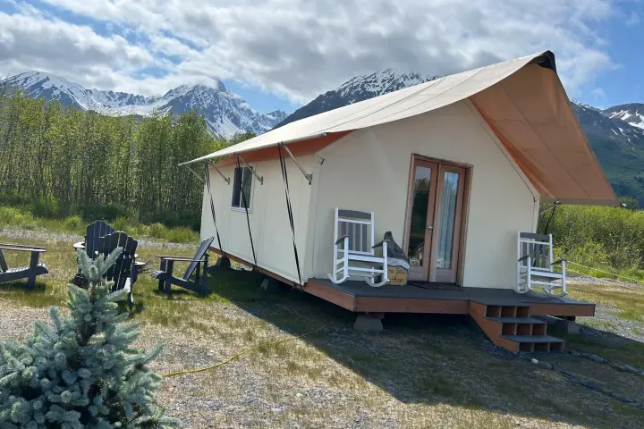 Tent cabin with chairs on porch, in mountainous landscape with snow-capped peaks and greenery.