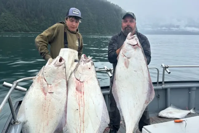 Two people on a boat holding large fish against a forested mountain backdrop.