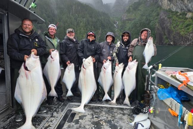 Group of seven people on a boat holding large fish, with a forested mountain in the background.