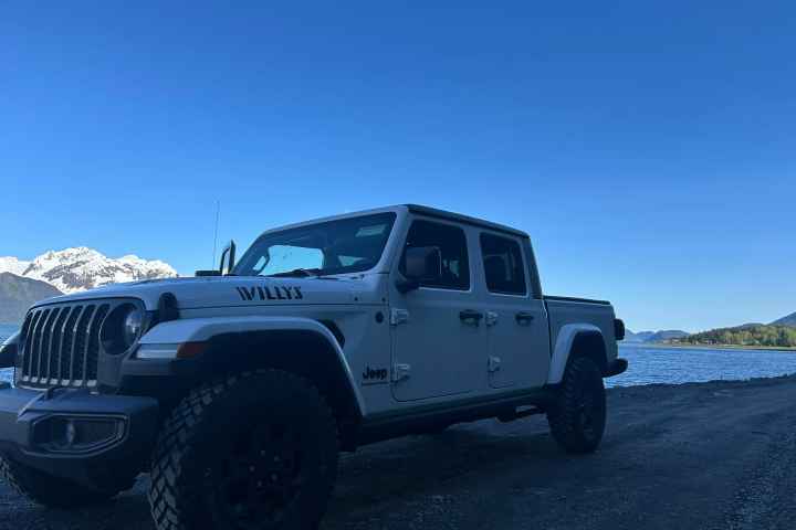 White Jeep parked by a lake with snow-capped mountains under clear blue sky.