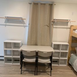 Room with two chairs at a desk, shelves, and beige curtains on a wood floor.