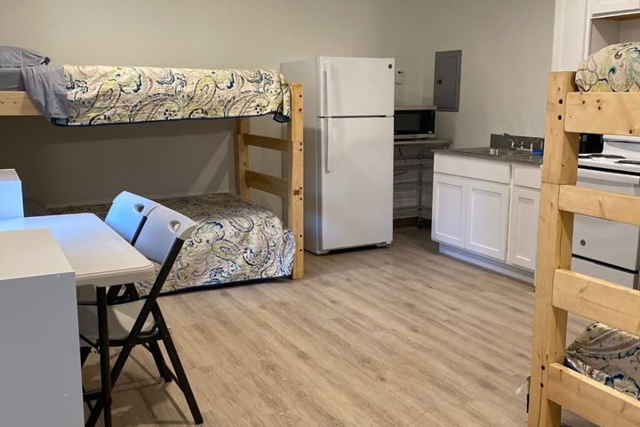 Room with bunk beds, fridge, microwave, table, and folding chairs on wood floor.
