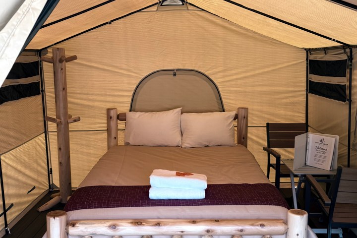 Interior of a canvas tent with a wooden bed, table, and chairs.