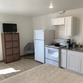 Small kitchen with fridge, stove, and sink, adjacent to a neatly arranged bed and bathroom.