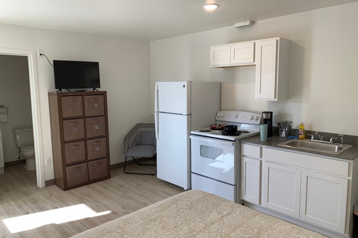 Small kitchen with fridge, stove, and sink, adjacent to a neatly arranged bed and bathroom.