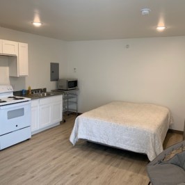 Studio apartment with a bed, kitchenette, fridge, and chair.