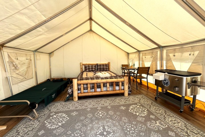 Inside a glamping tent with bed, table, chairs, cot, and cooler on a patterned rug.