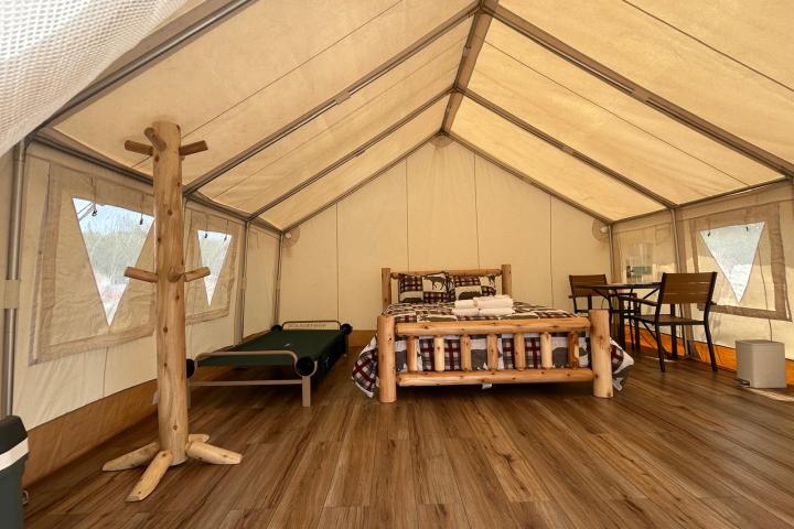 Inside view of a glamping tent with a wooden bed, side table, and chairs on a wooden floor.