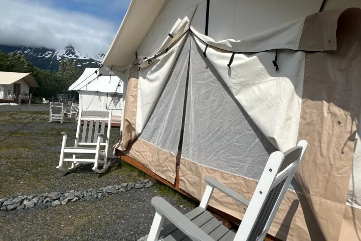 Campsite with large canvas tents and white rocking chairs under a blue sky.