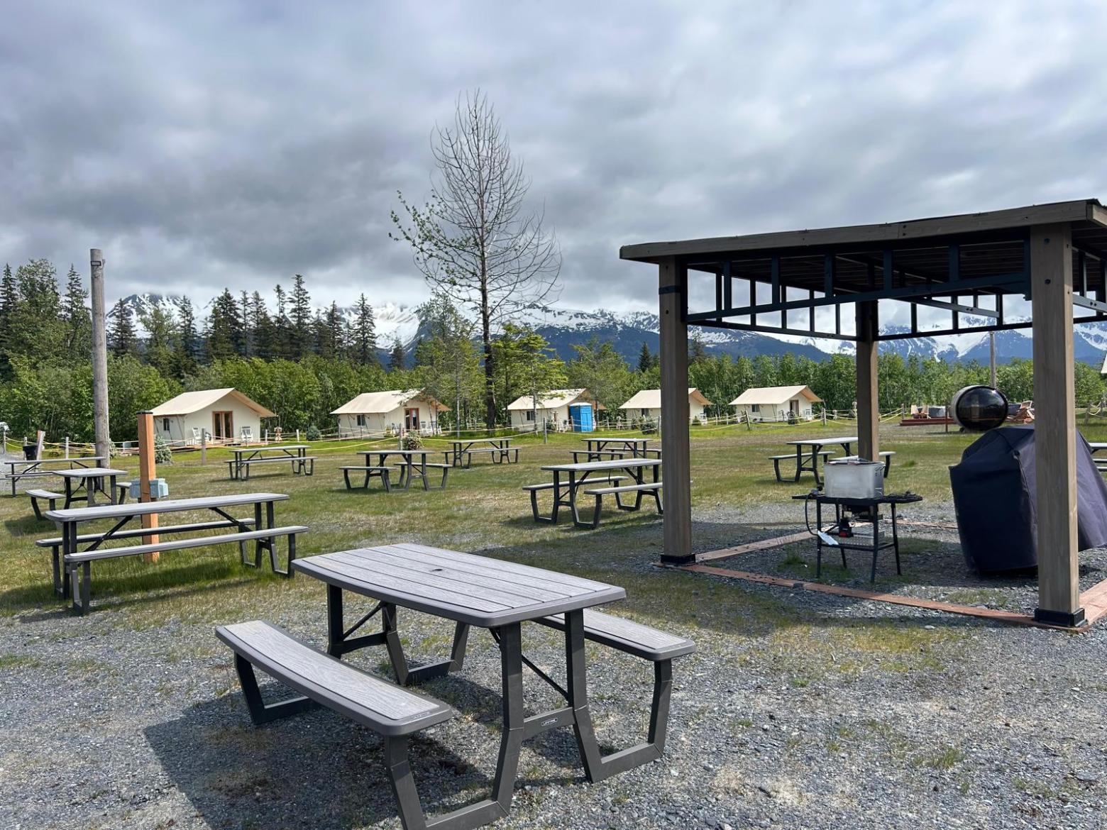 Outdoor campsite with picnic tables, tents, and a grill under a cloudy sky.