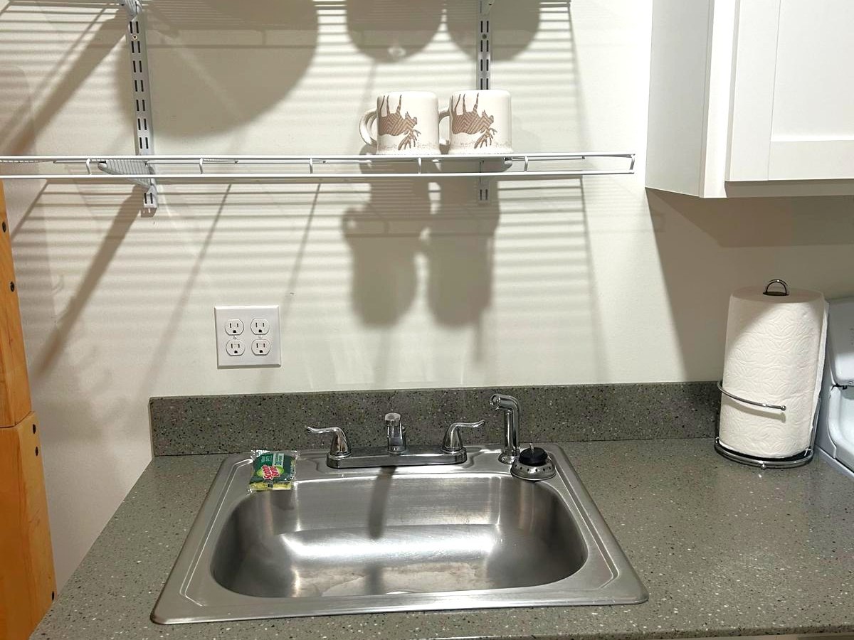 Kitchen sink with gray countertop, shelves with dishes, and a paper towel holder.