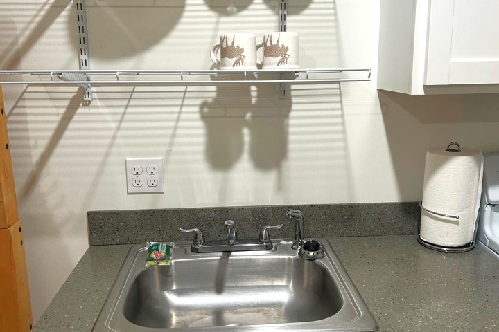 Kitchen sink with shelves above holding dishes and mugs, paper towel on counter.