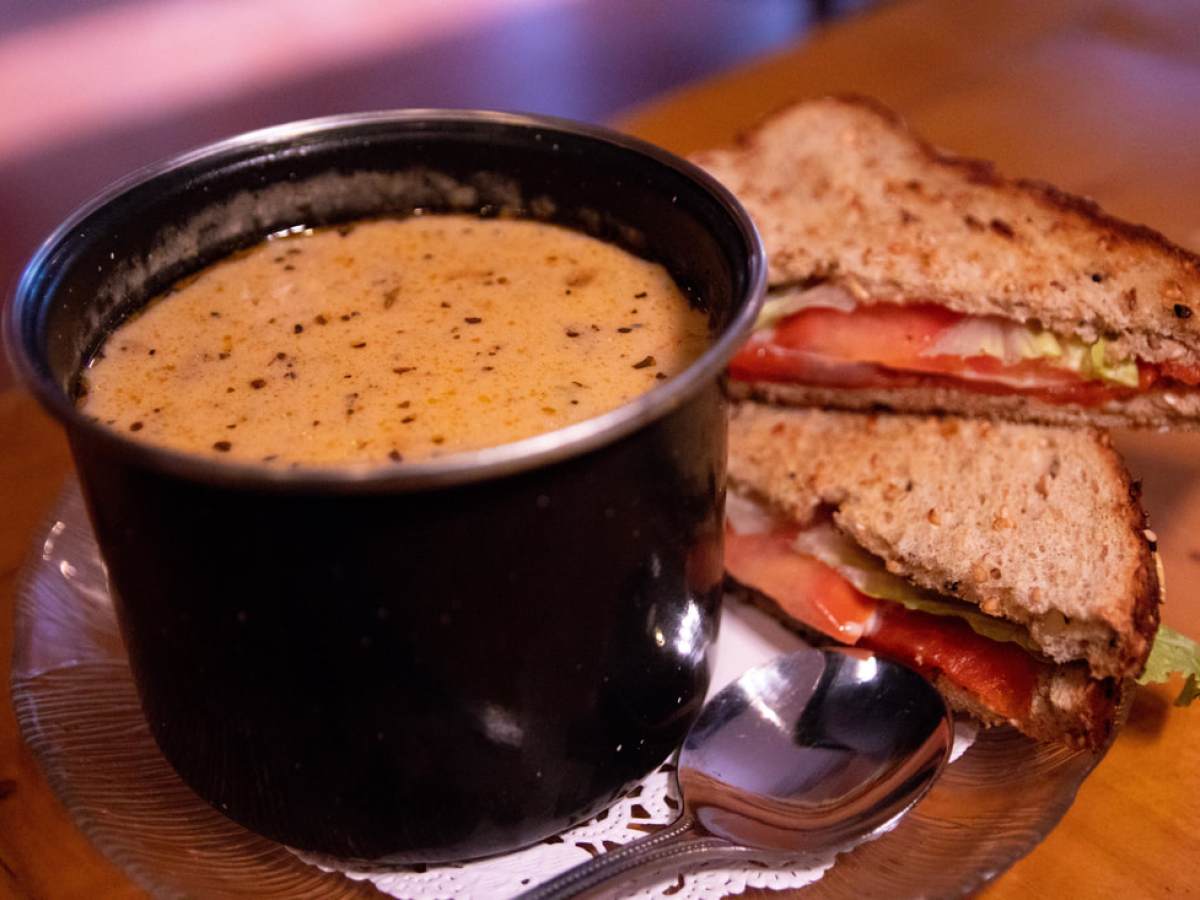 Bowl of soup with pepper beside a sandwich on a plate with a spoon.