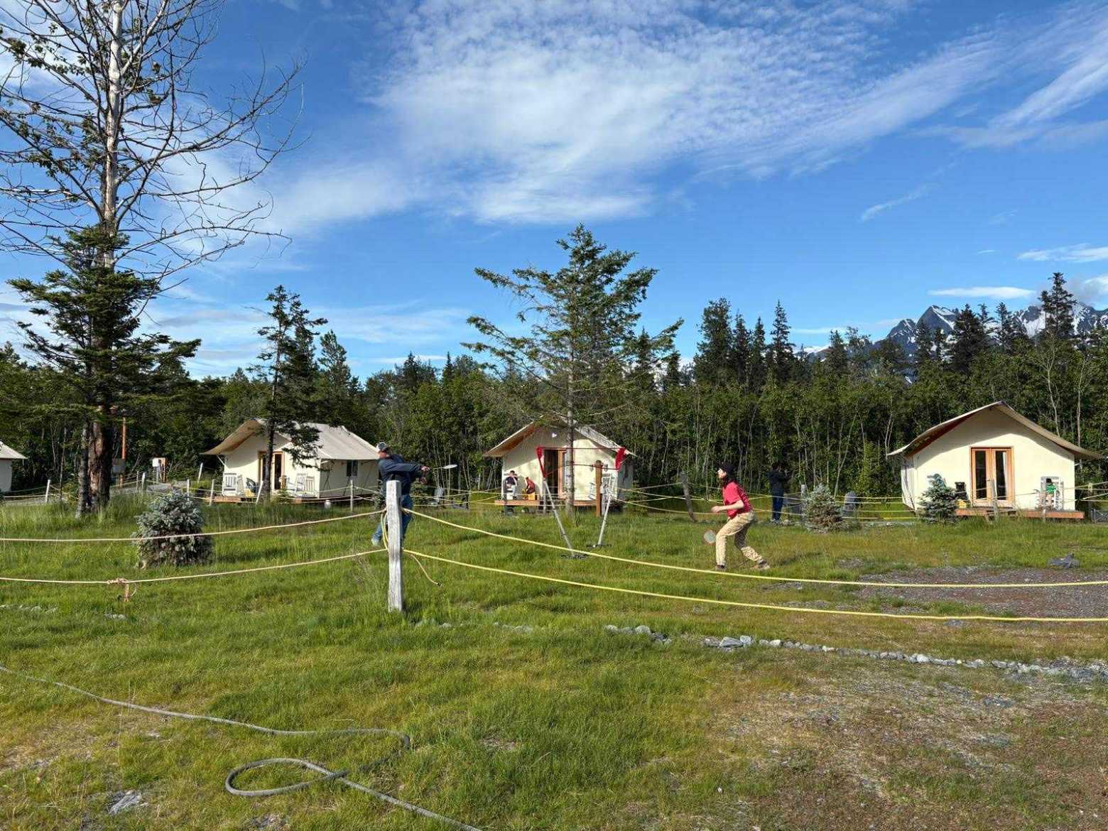 People playing badminton on grassy area with small cabins and trees.
