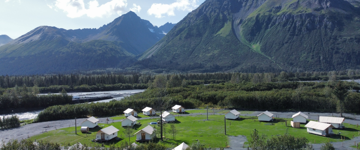 Aerial view of cabins on green grass with mountains in the background.