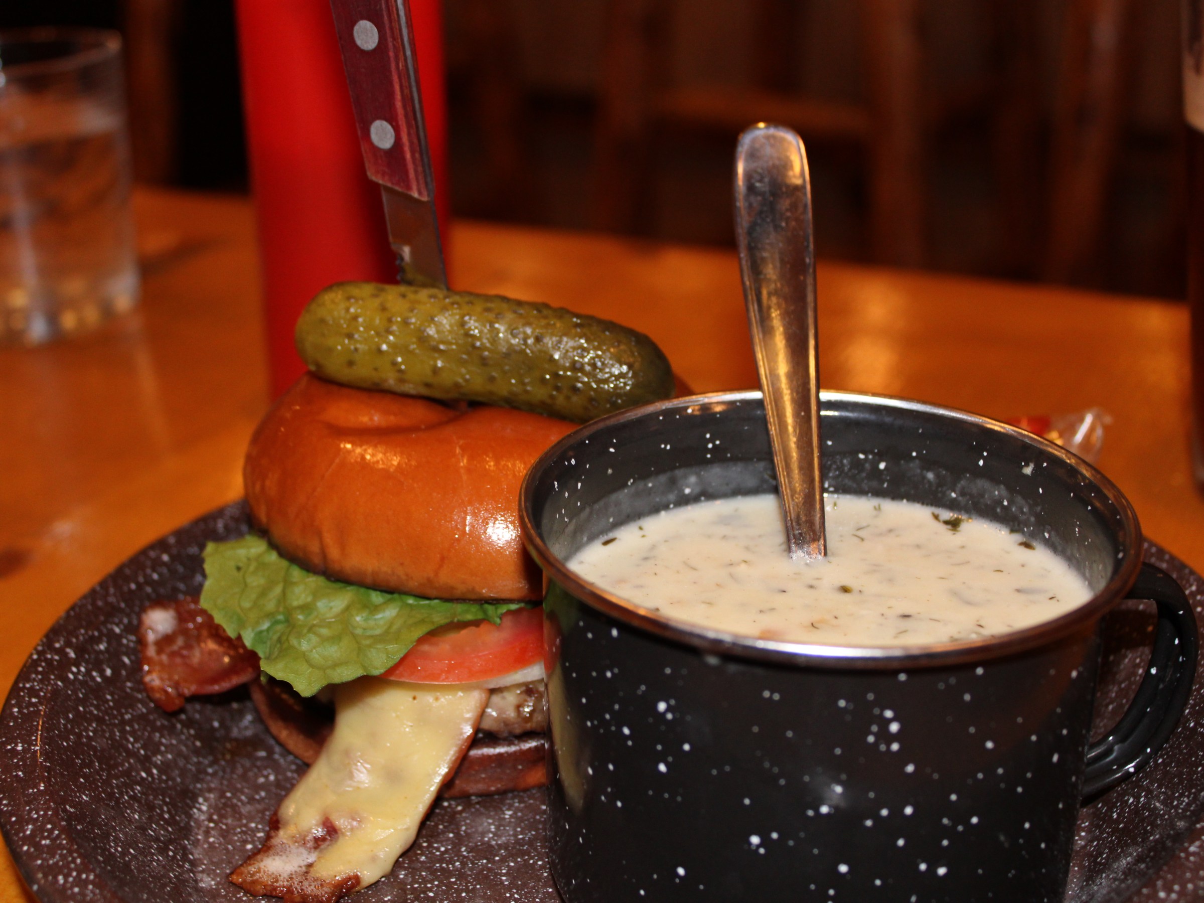 Burger with pickle and knife next to a mug of creamy soup on a wooden table.