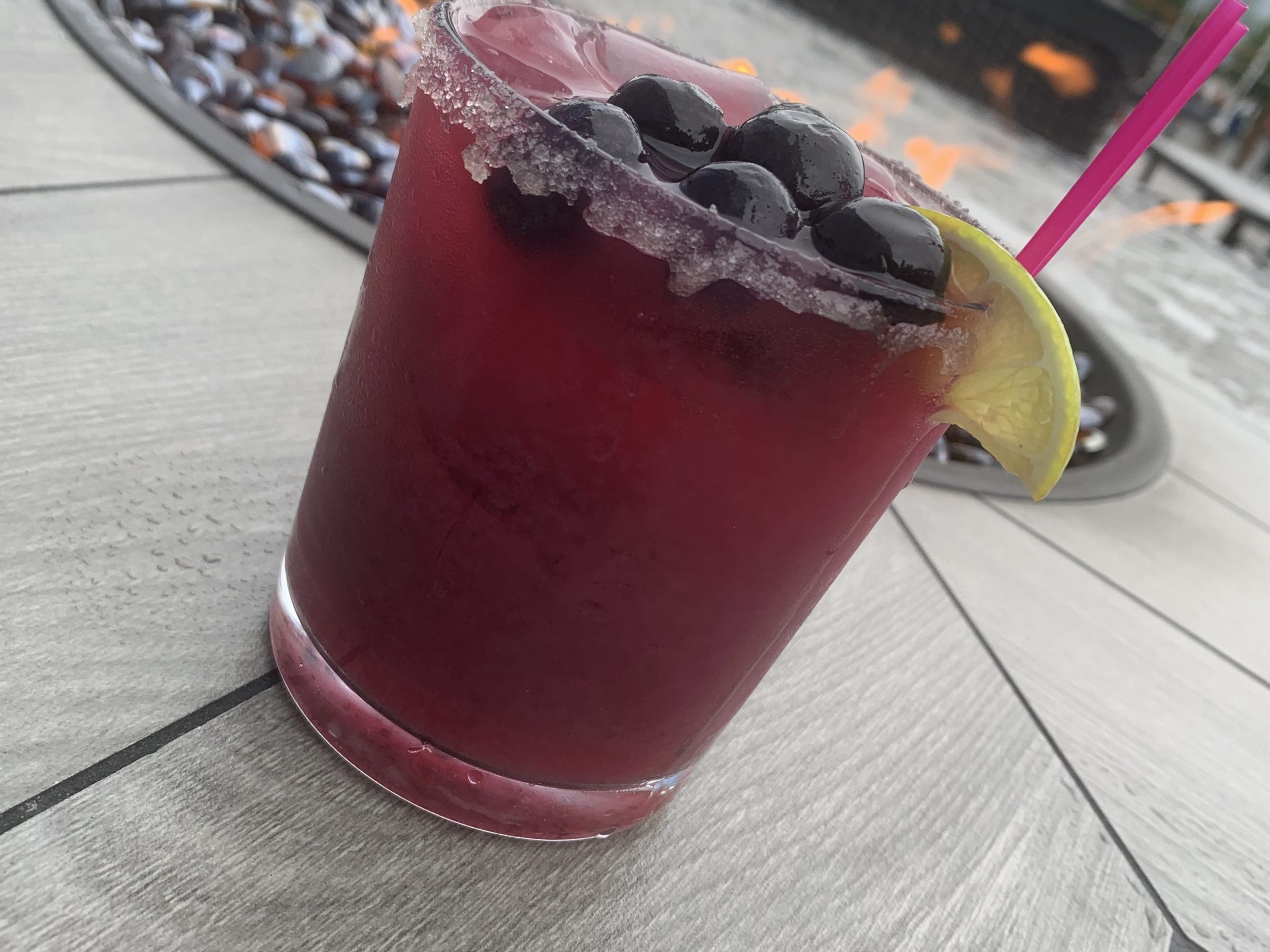 Purple cocktail with blueberries, lemon slice, pink straw by outdoor fire pit.