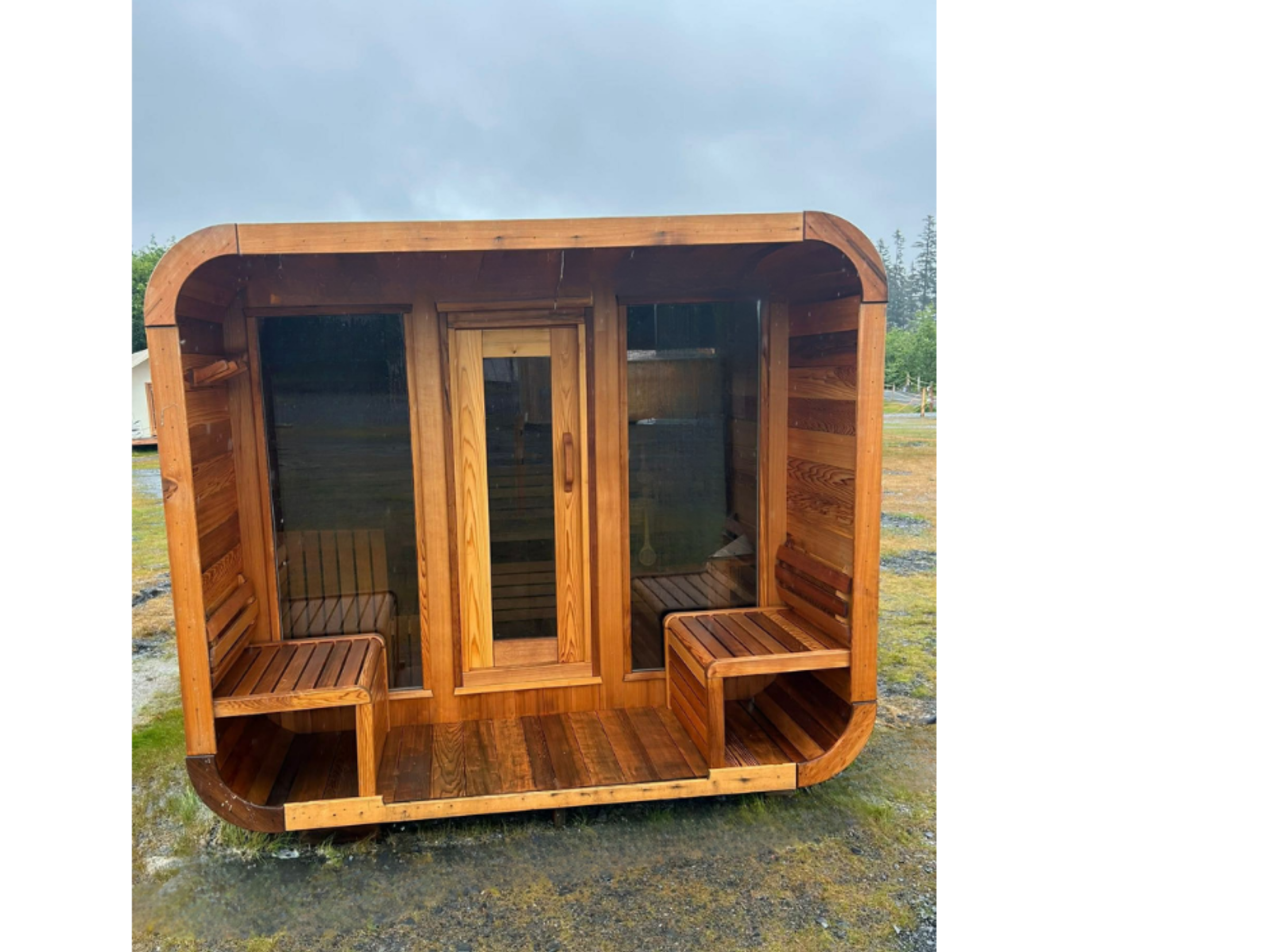Wooden sauna with glass doors and bench seats, outdoors on a rainy day.