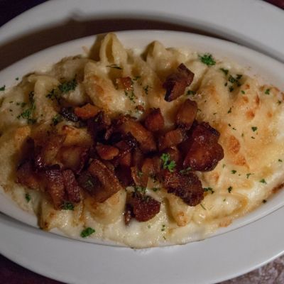 Baked mac and cheese topped with crispy bacon pieces in a white dish on a plate.
