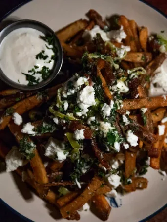 Plate of fries with cheese, herbs, and dip on the side on a wooden table.
