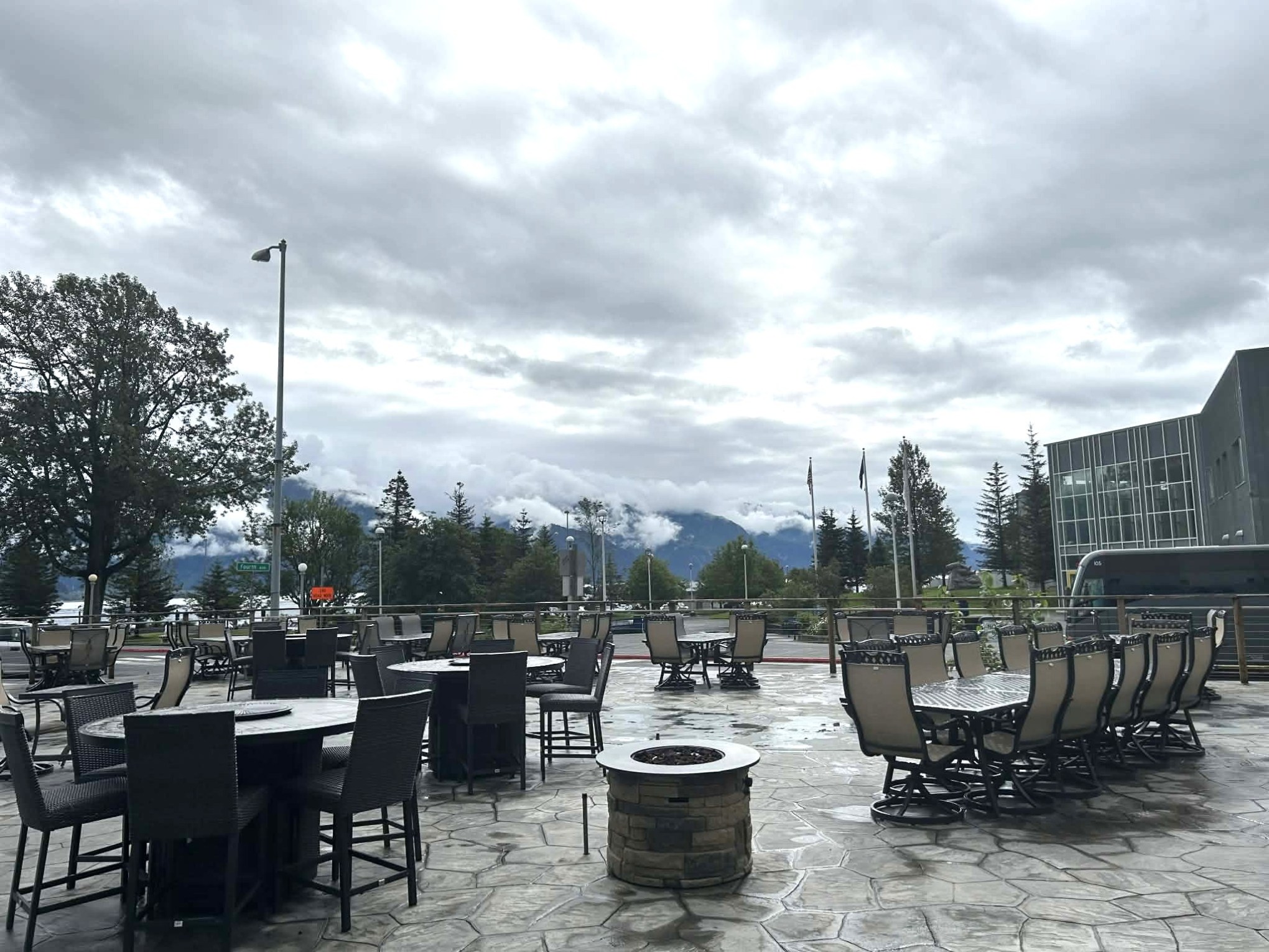 Outdoor patio with empty tables and chairs, cloudy sky, and trees in the background.