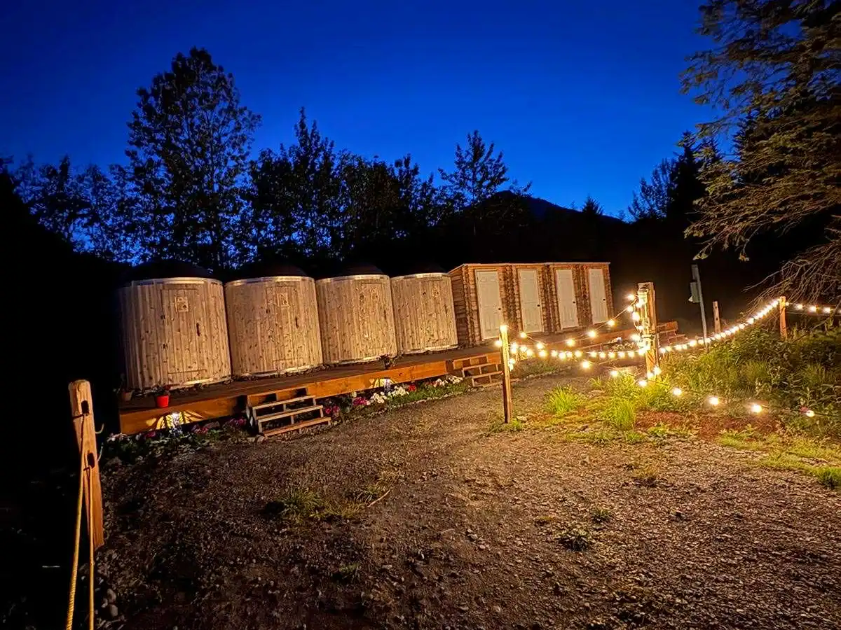 Outdoor wooden cabins with string lights at night, framed by trees.