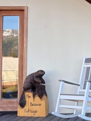 Wooden beaver sculpture beside rocking chair near a door labeled 'The Beaver Cottage' on a porch.