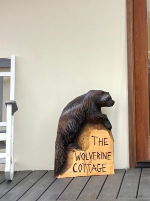 A wooden wolverine sculpture on a porch beside a sign reading 'The Wolverine Cottage'.
