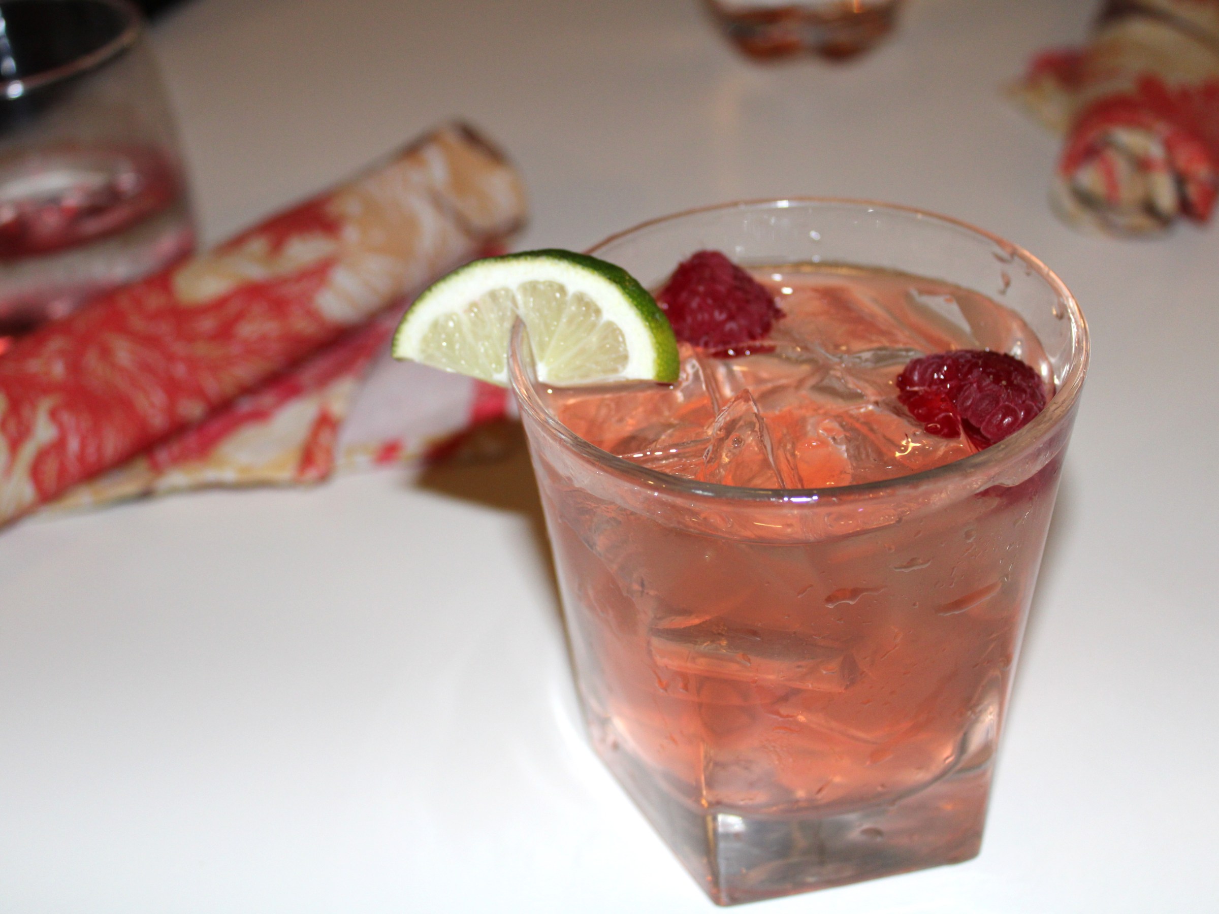 Pink cocktail with ice, raspberries, and lime slice in a glass on a table.