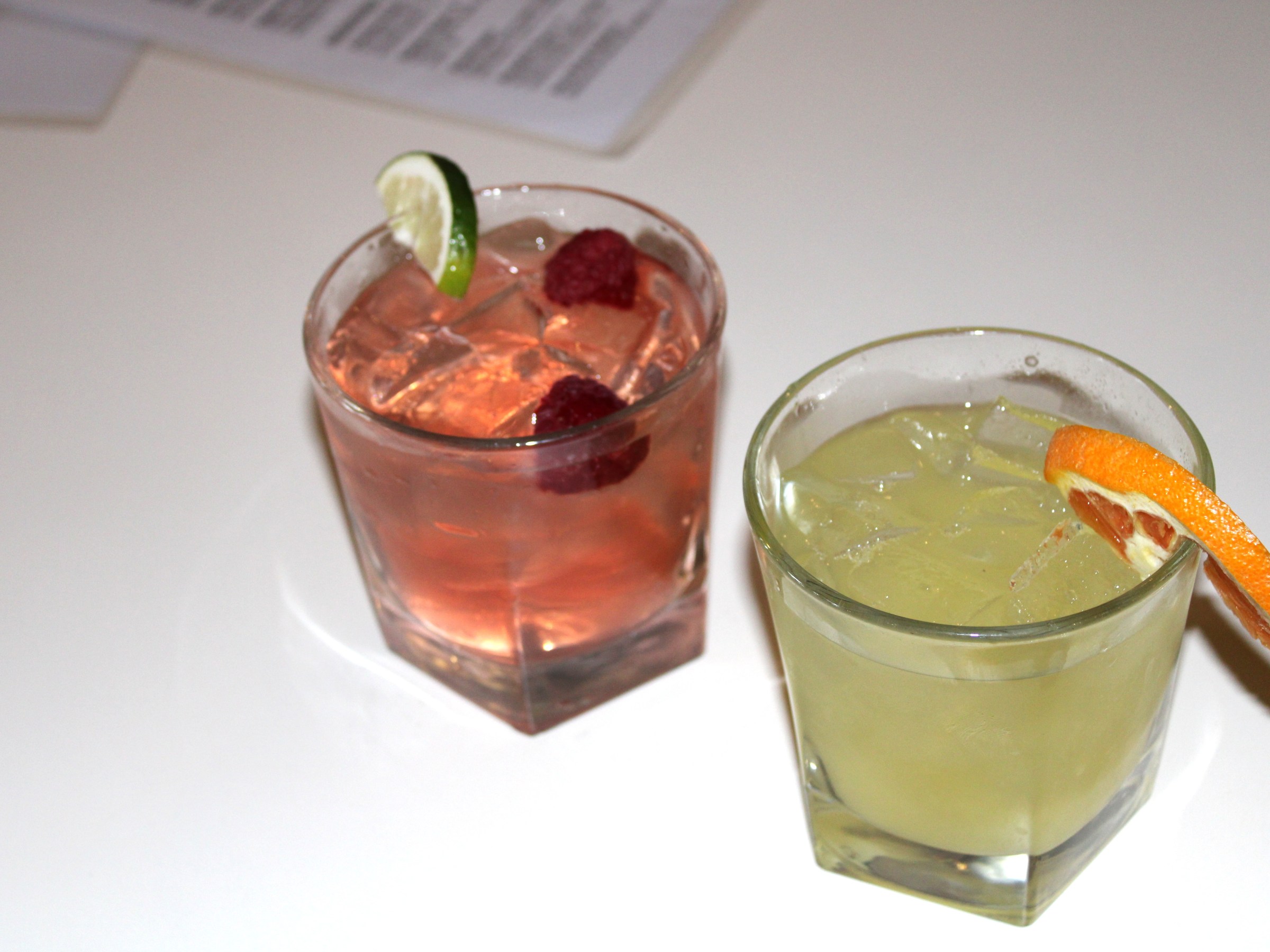 Two cocktails on a table, one pink with raspberries and lime, the other yellow with an orange slice.