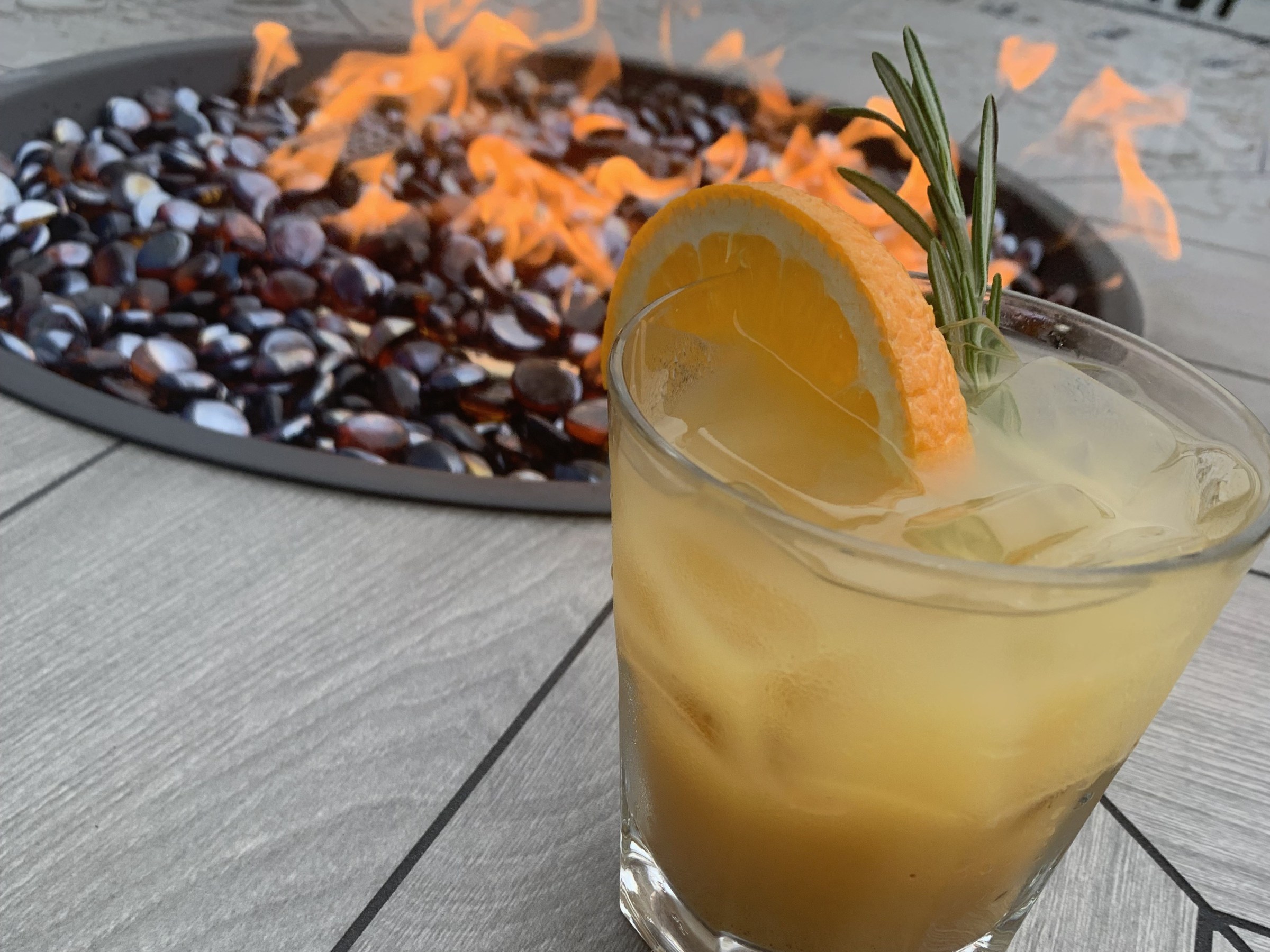 Cocktail with orange slice and rosemary by a fire pit on a wooden table.