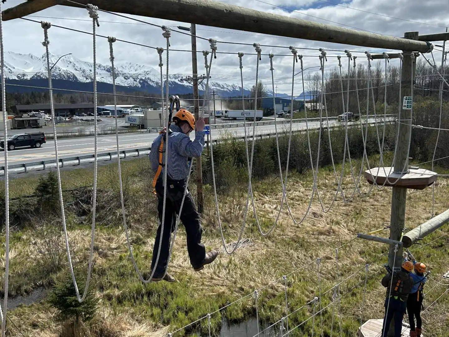 Person on ropes course with helmet, mountains and cloudy sky in background.