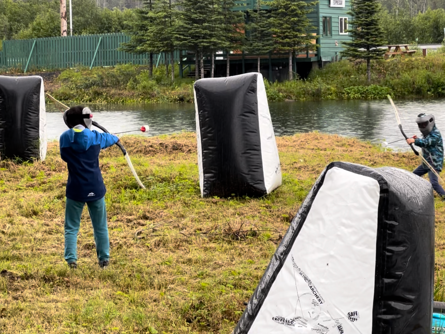 Two people in gear playing combat archery near inflatable obstacles by a river.