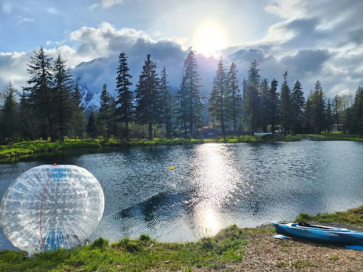 Lake with a transparent inflatable ball and kayak, surrounded by trees and mountains under a bright sun.