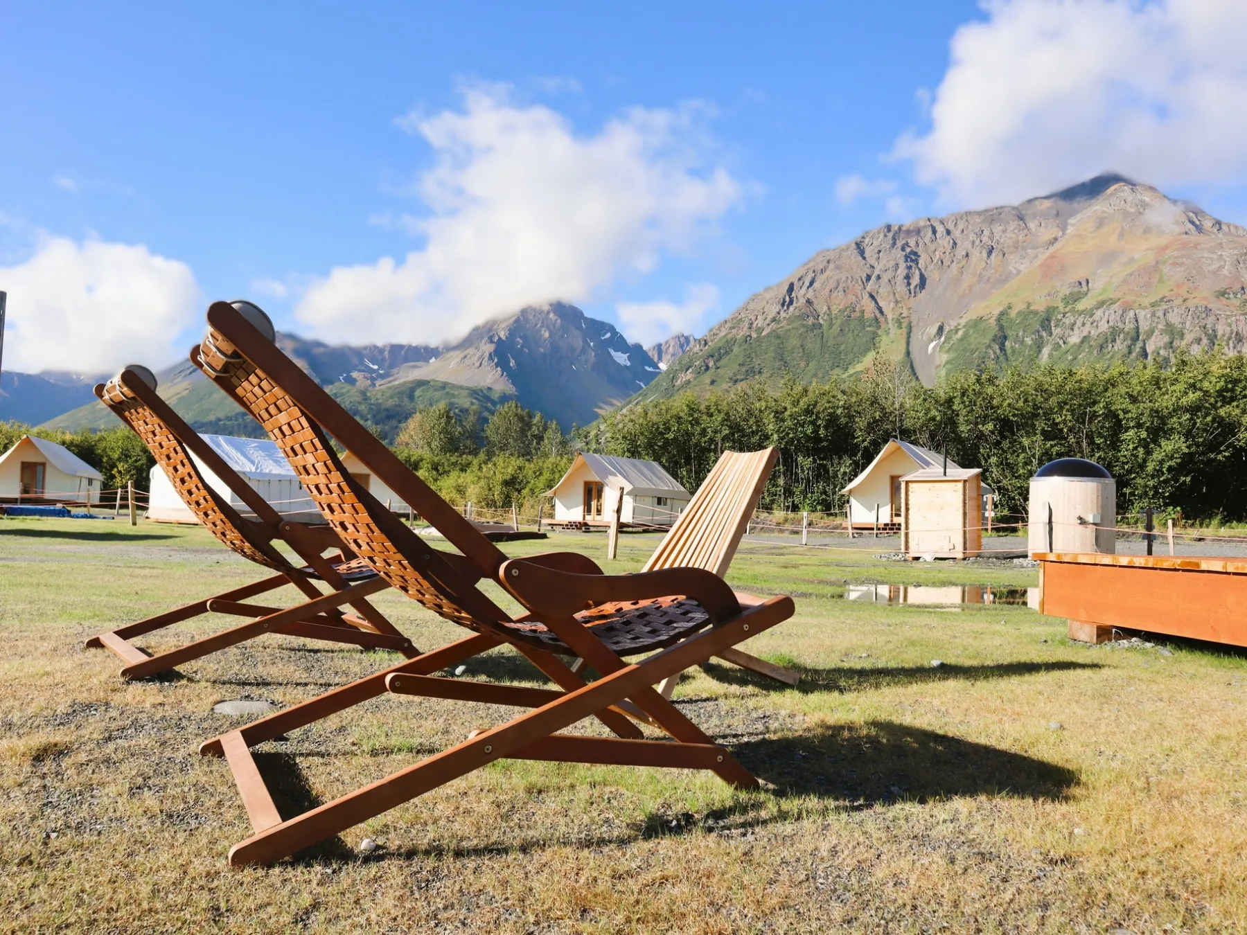 Wooden lounge chairs on grass with mountains and tents in the background.