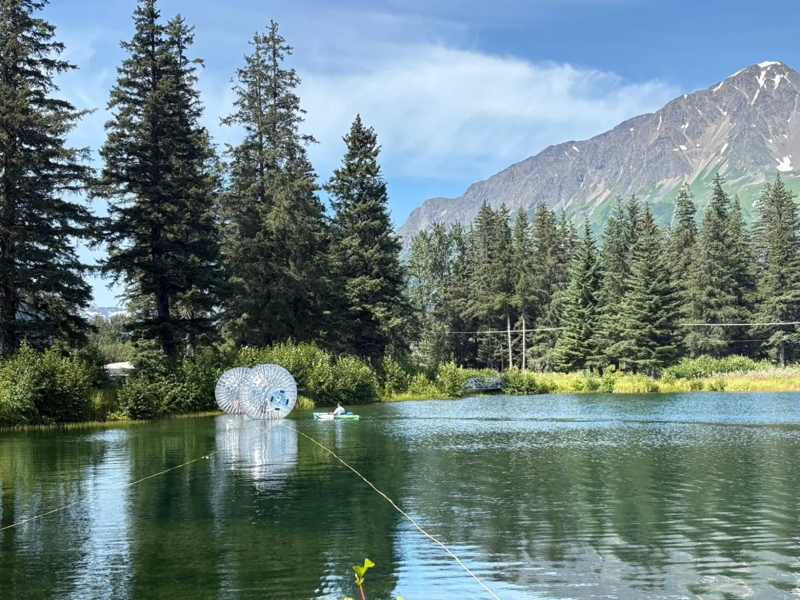 Lake with a zorb and a kayak, surrounded by trees and mountains under a clear blue sky.