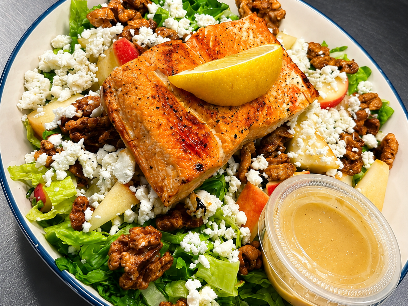 Salad with grilled salmon, walnuts, feta, apple slices, and dressing cup on side.